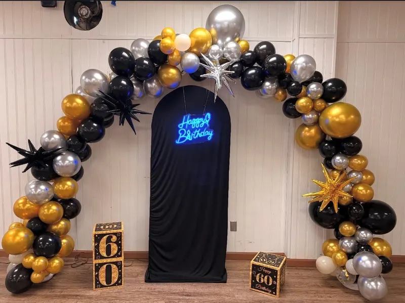 A black, gold and silver balloon arch with starburst accents around a black drape and Happy Birthday neon sign, with printed 60 gift boxes at the base.