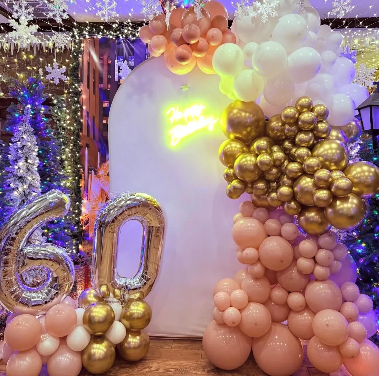 A blush, white and gold balloon garland on a white arch backdrop with a Happy Birthday neon sign and silver 60 balloons.