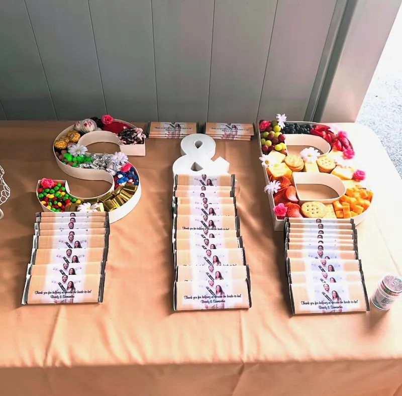 Letter-shaped trays spelling S and B filled with candy and charcuterie beside rows of personalized photo chocolate bar favors on a peach-linen table for a bridal shower.