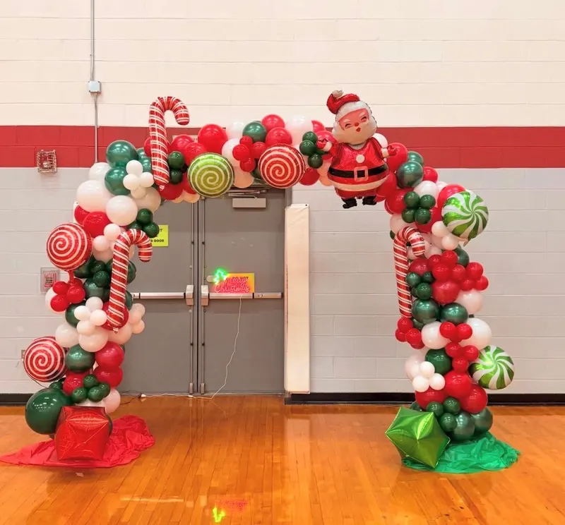 A red, green and white balloon arch built with candy cane and peppermint swirl balloons and a Santa foil balloon, framing a gymnasium doorway.