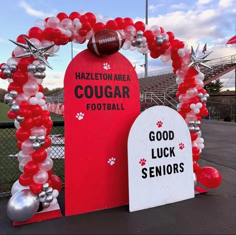 A red, white and silver balloon arch with a foil football and starburst balloons framing Hazleton Area Cougar Football and Good Luck Seniors backdrop signs at a football field.