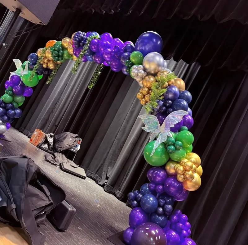 A jewel-toned balloon arch in purple, green, gold and silver accented with iridescent fairy wings and greenery, set on a dark stage.