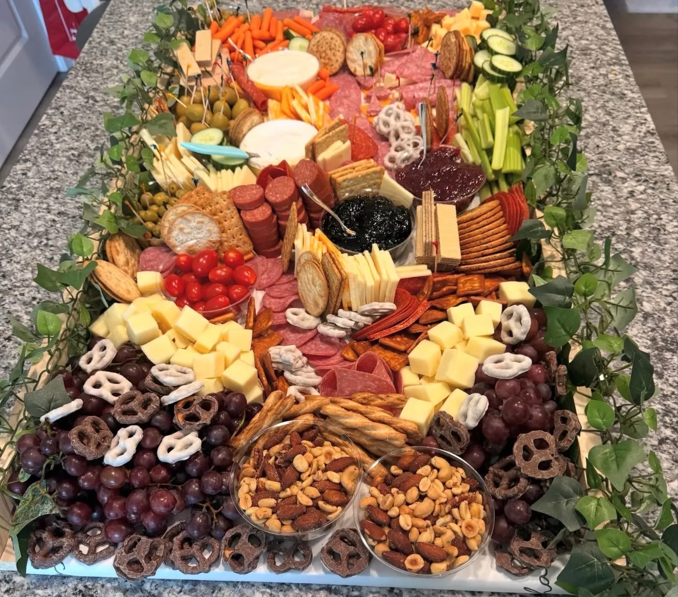 A rectangular grazing spread of cheeses, cured meats, fruit, nuts, and pretzels edged with greenery on a granite counter.