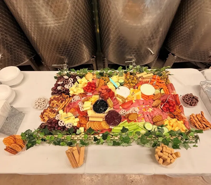 A large square grazing table covered edge to edge with cheeses, cured meats, crackers, fruit and dips, bordered with fresh greenery, set in front of stainless steel winery tanks.
