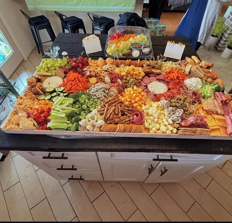 A generous grazing board covering a rustic wood plank across a kitchen island, with vegetables, cheeses, crackers and dips arranged in bright color-blocked rows.