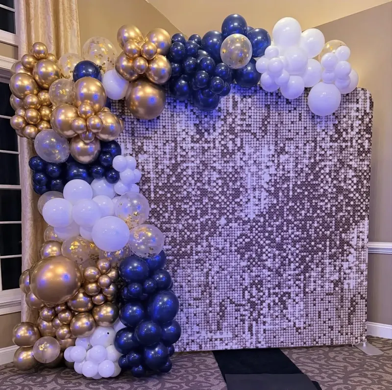A navy, gold and white balloon garland cascading down the side of a rose-gold shimmer sequin wall in an elegant carpeted room.