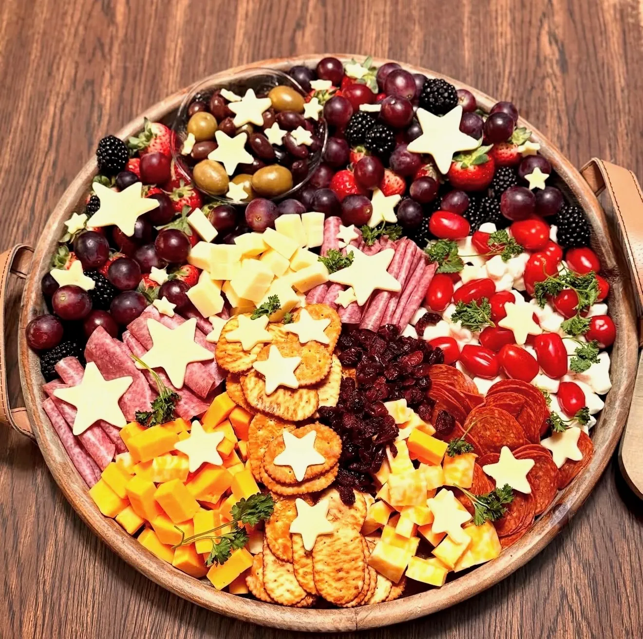 A round charcuterie board with red fruit, dark grapes, meats, crackers, and star-cut cheeses.
