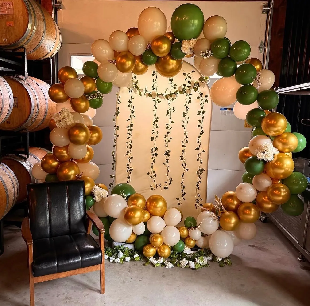 A circular balloon ring in sage green, cream and gold with trailing greenery and a fabric backdrop, set among oak wine barrels beside a leather chair.