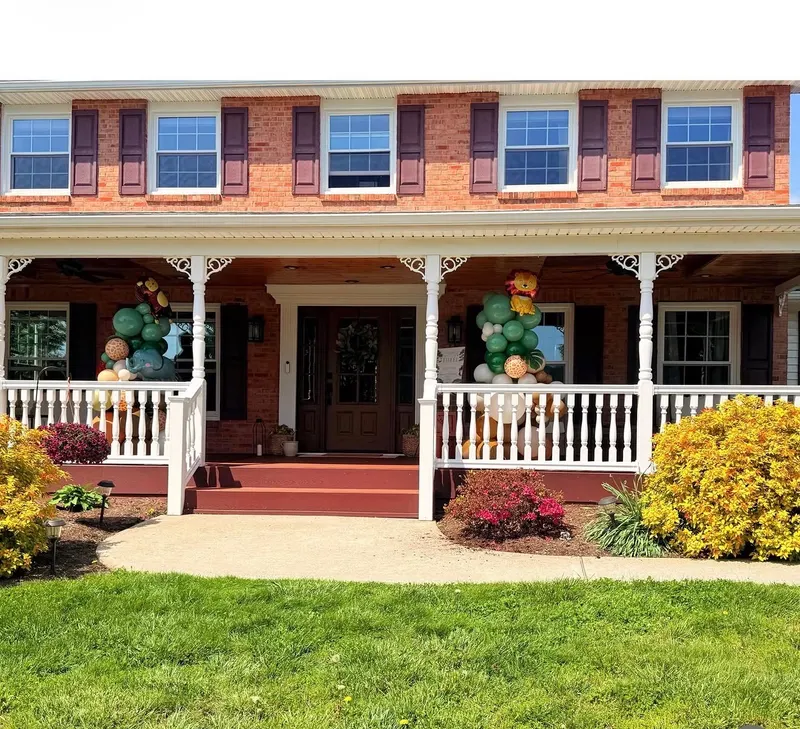A pair of green, tan and honey-gold balloon columns topped with Winnie the Pooh characters, flanking the front door of a brick home with a covered porch.